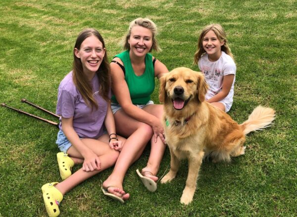 A golden retriever sitting on the grass with a mum and her two daughters, all smiling up at the camera